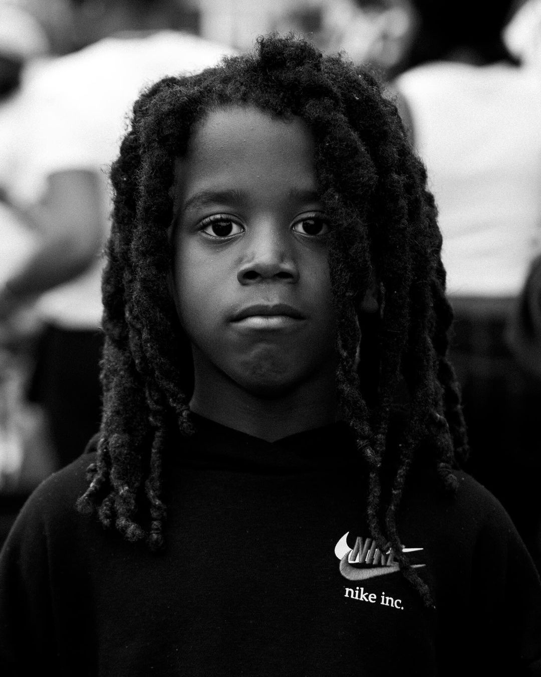 Portrait of a young Black child with dreadlocks, wearing a black Nike sweatshirt, photographed in black and white with an outdoor background.