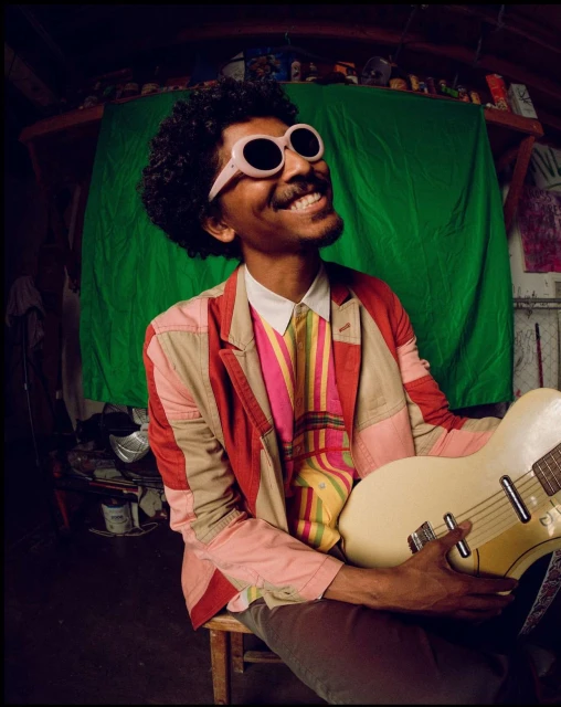 Smiling man in sunglasses and a colorful blazer holds a guitar in an indoor space with a green screen and various objects.