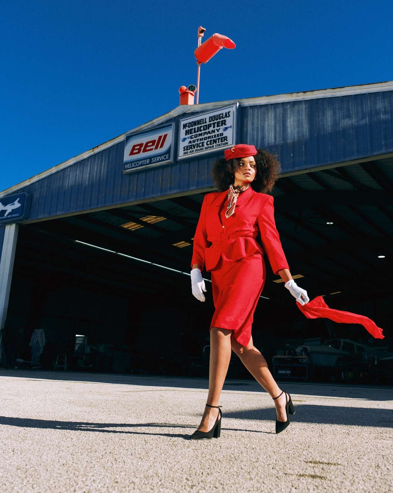 A woman in a red suit walks in front of an aircraft hangar under a clear blue sky, creating a striking and vintage-style fashion image.