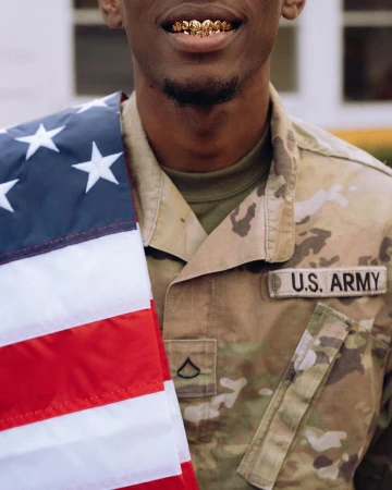 Man in U.S. Army uniform with gold teeth grill, holding a portion of the American flag in an indoor setting.