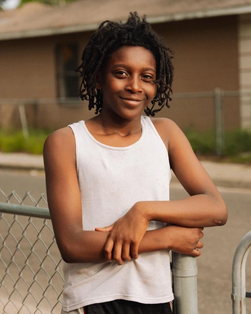 Smiling young Black woman with dreadlocks, wearing a white tank top, leans against a grey metal fence in front of a house.