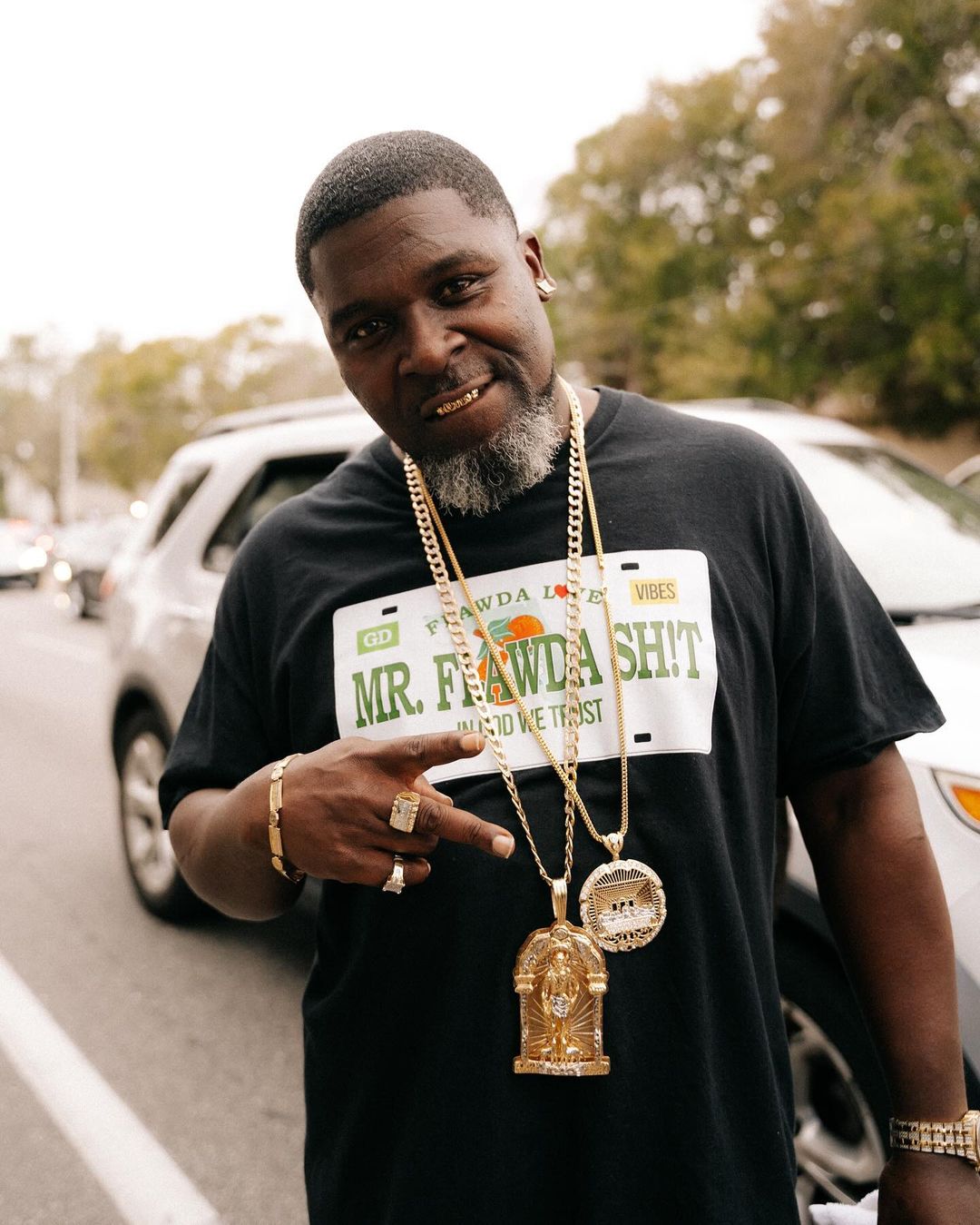 Portrait of a Black adult male wearing gold jewelry and a black graphic t-shirt outdoors against an urban street backdrop with cars and trees.