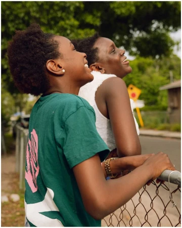 Two African-American women laugh together outdoors by a chain-link fence, wearing casual clothing with a green shirt and white top, shot in daylight.