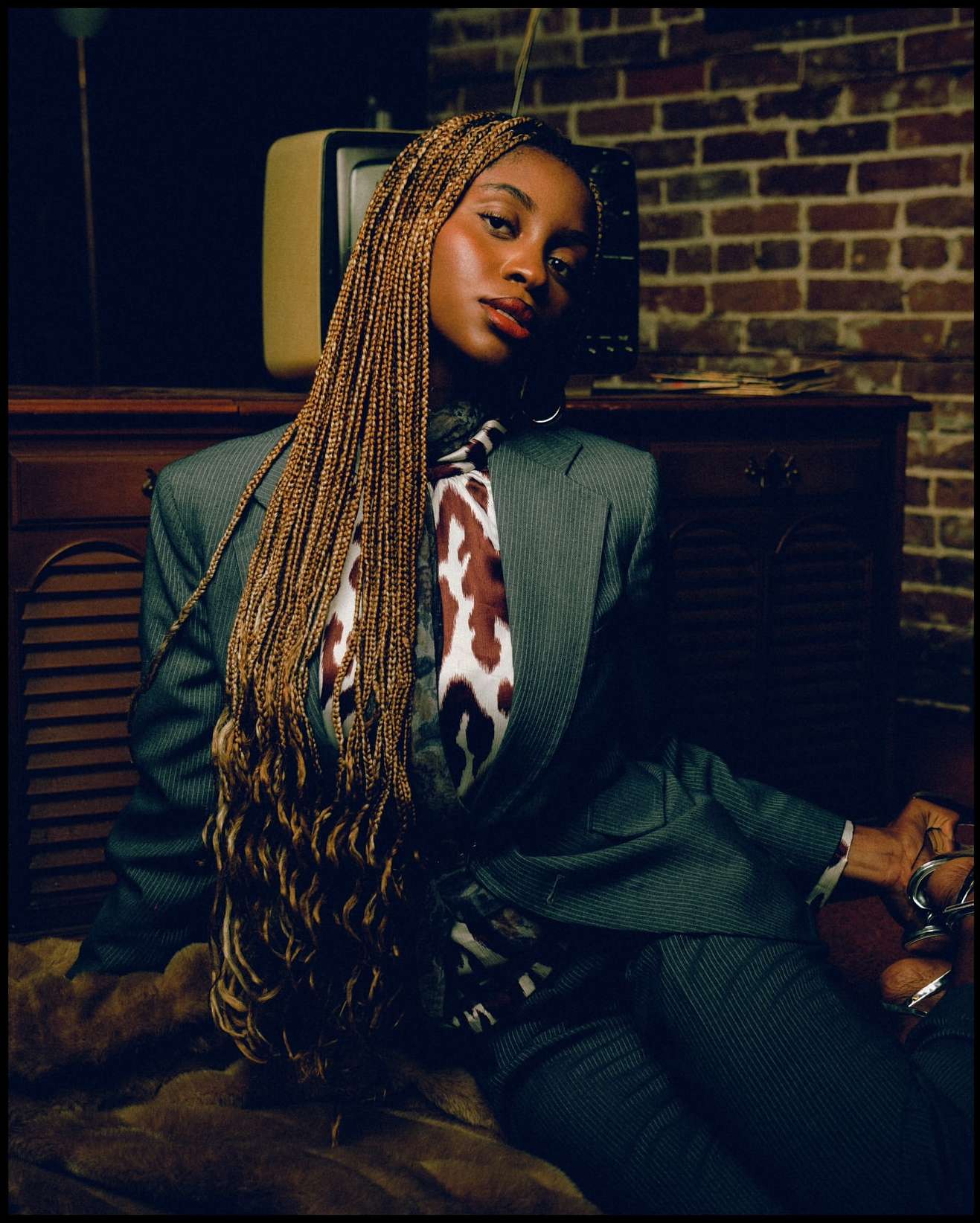 Portrait of a stylish African-American woman in a gray suit, posing indoors with long braids against a brick wall and vintage TV.