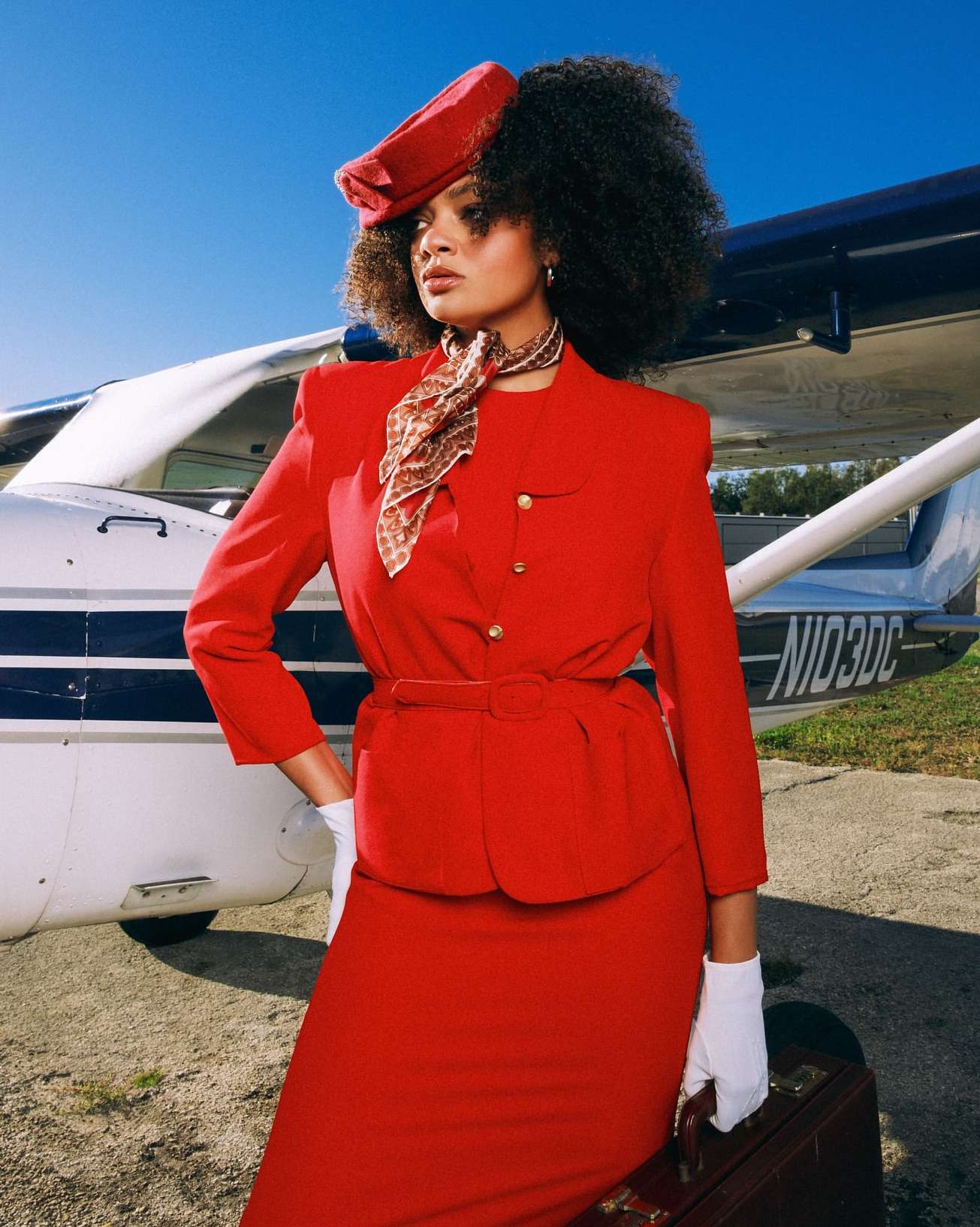An adult woman poses in front of a small airplane wearing a red skirt suit, matching hat, and white gloves, carrying a brown suitcase.