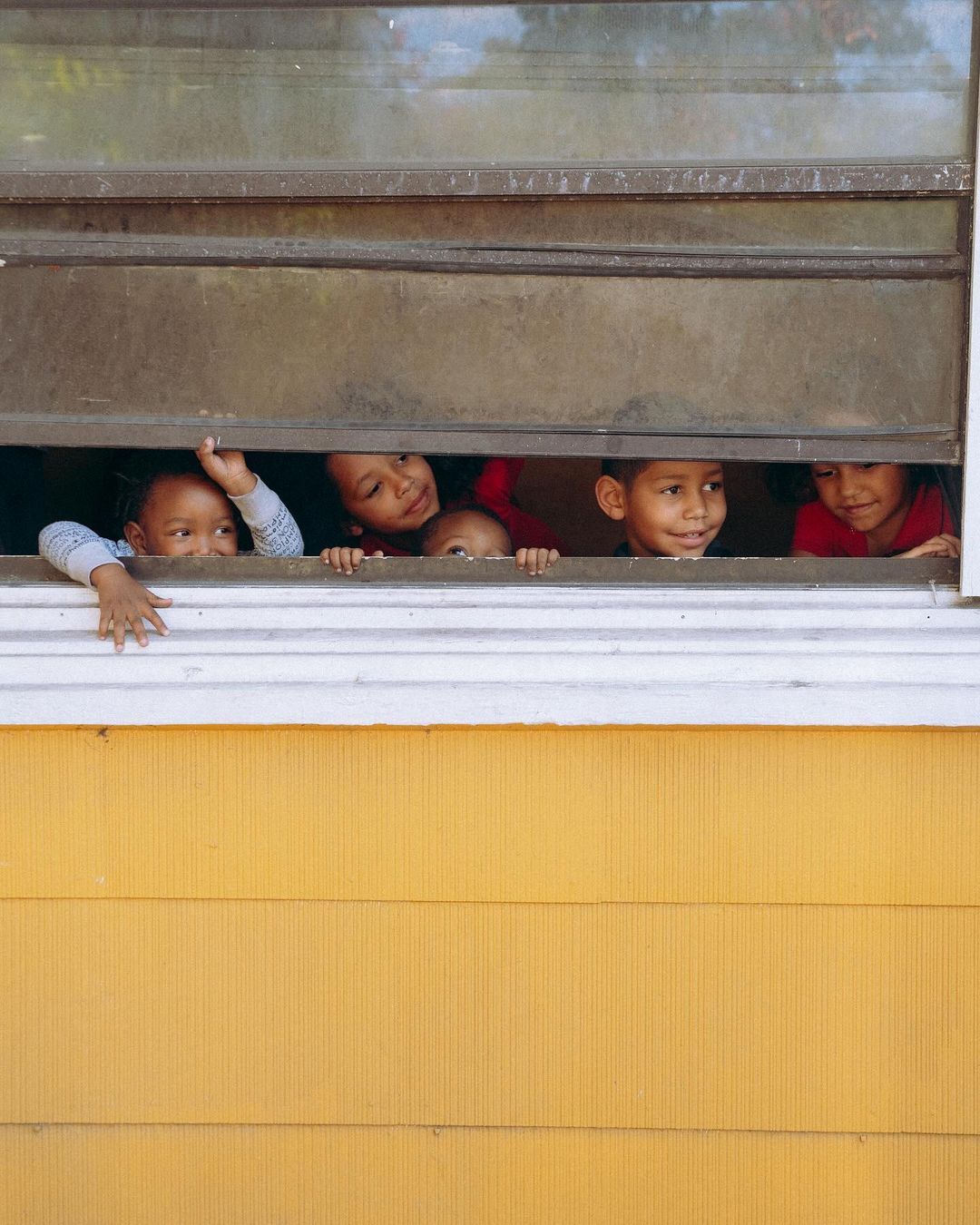 Five smiling children peer through a window with closed blinds, their faces lit by soft light and backed by a yellow building exterior.