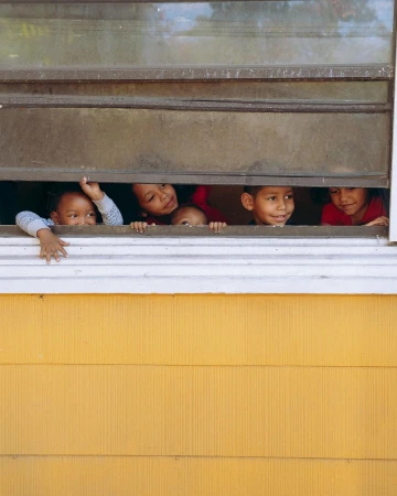 Five smiling children peer through a window with closed blinds, their faces lit by soft light and backed by a yellow building exterior.