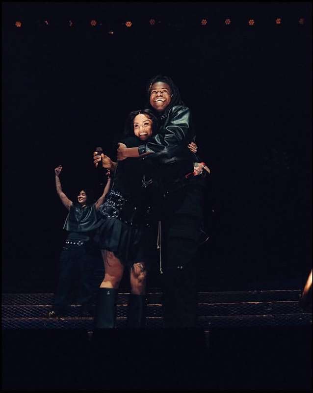 Two singers hug on stage during a performance, bathed in stage lighting, as a third performer stands with arms raised in the background.