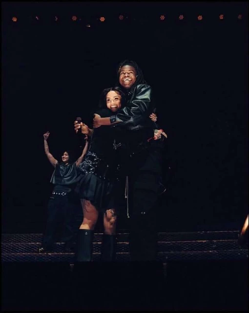 Two singers hug on stage during a performance, bathed in stage lighting, as a third performer stands with arms raised in the background.