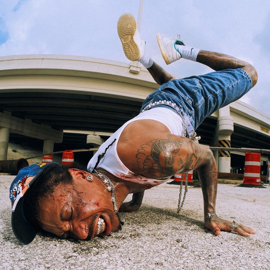 Adult male breakdancer on concrete surface wearing a white tank top, denim shorts, sneakers, and cap; urban environment and bright daylight.