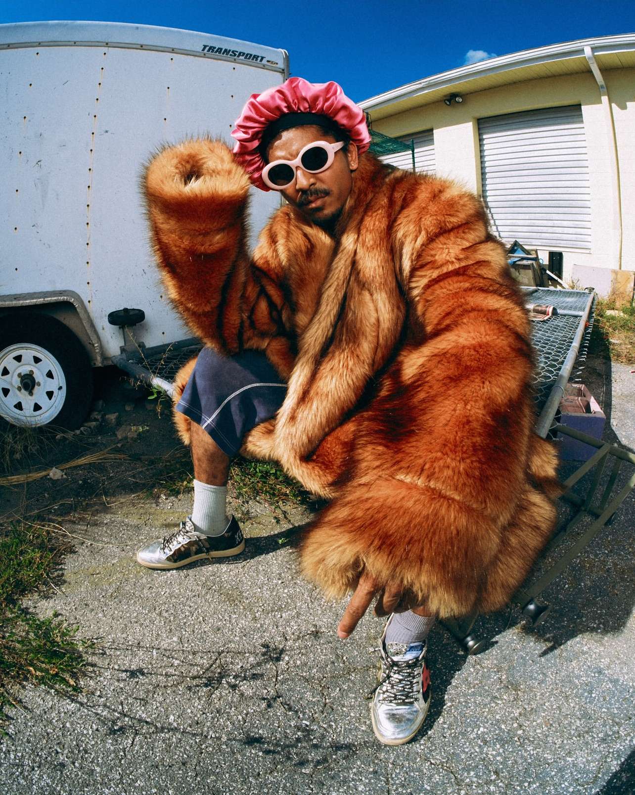 A man wearing a fur coat, pink bonnet, and sunglasses sits outdoors by a trailer. He is wearing silver sneakers and a pointed index finger.