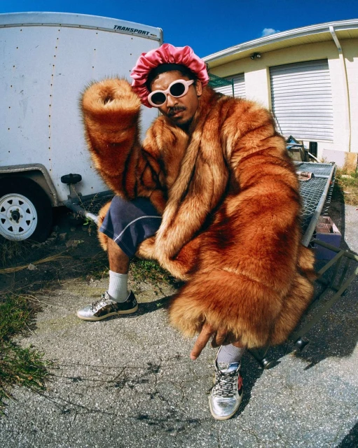 A man wearing a fur coat, pink bonnet, and sunglasses sits outdoors by a trailer. He is wearing silver sneakers and a pointed index finger.