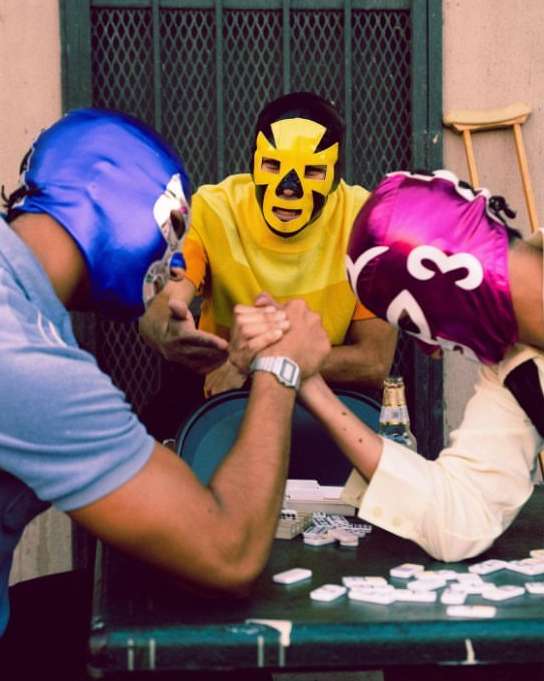 Three masked wrestlers engage in arm wrestling at a table outdoors, surrounded by dominoes and a beer bottle in a gritty scene.
