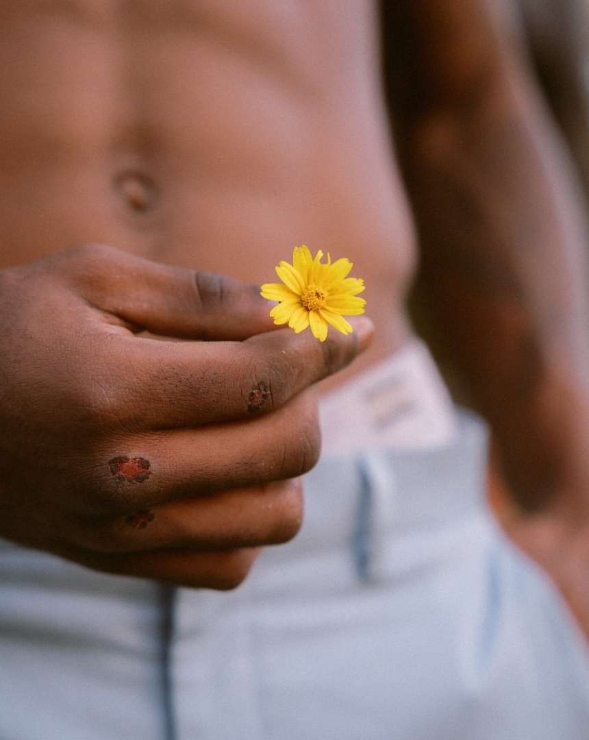 Close-up of a tan-skinned torso and hand gently holding a small yellow flower. Light blue denim pants are visible at the waist.