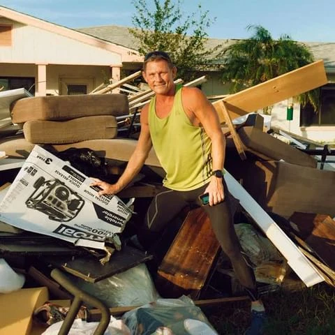 Adult male standing in front of a pile of debris and discarded household items after a flood or natural disaster, holding a generator box.