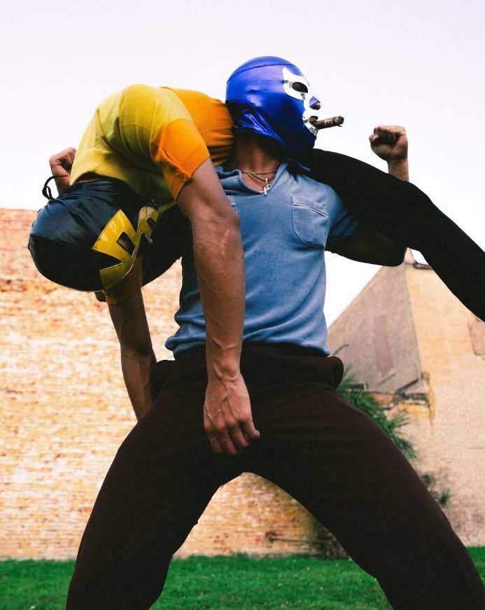 Two masked luchadores in a wrestling-inspired pose outdoors. One wrestler carries the other on his shoulders in a display of strength.
