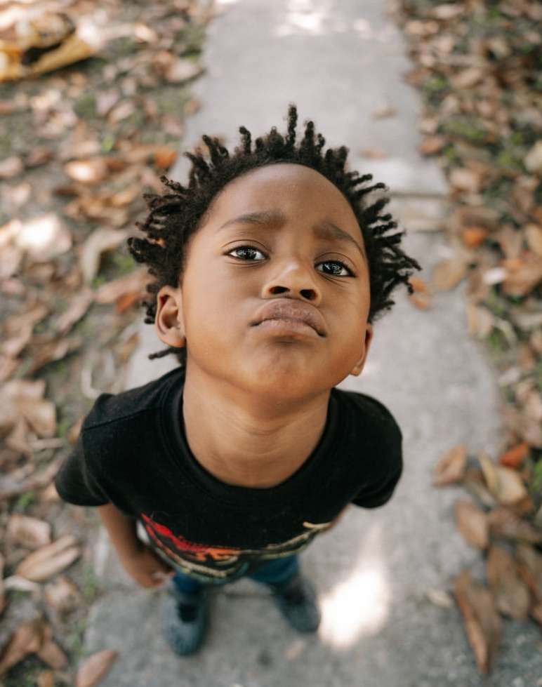 Young boy with dark hair looks up at the camera with a serious expression while standing on a sidewalk covered in fallen leaves.