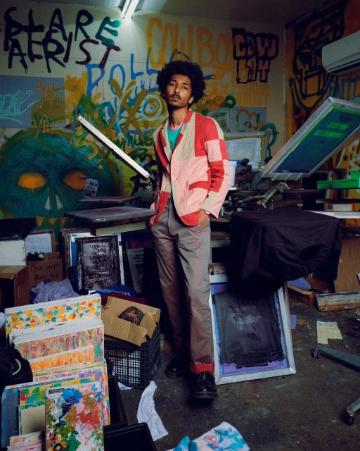 Adult man in a patchwork jacket poses in an indoor setting with artistic wall graffiti and scattered art supplies creating a creative ambiance.