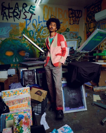 Adult man in a patchwork jacket poses in an indoor setting with artistic wall graffiti and scattered art supplies creating a creative ambiance.