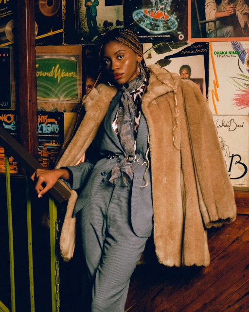 A stylish woman wearing a gray pinstripe suit and faux fur coat poses in front of vintage record album covers and a dark wooden railing.