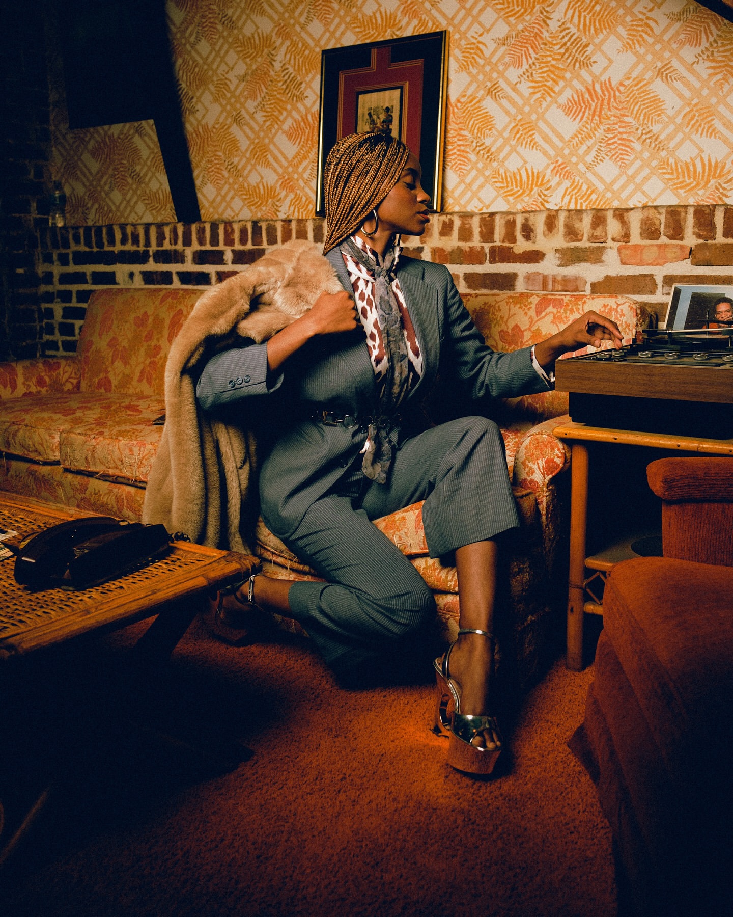 Stylish African-American woman in a suit and silver platforms poses indoors with a record player, fur coat, and vintage decor.