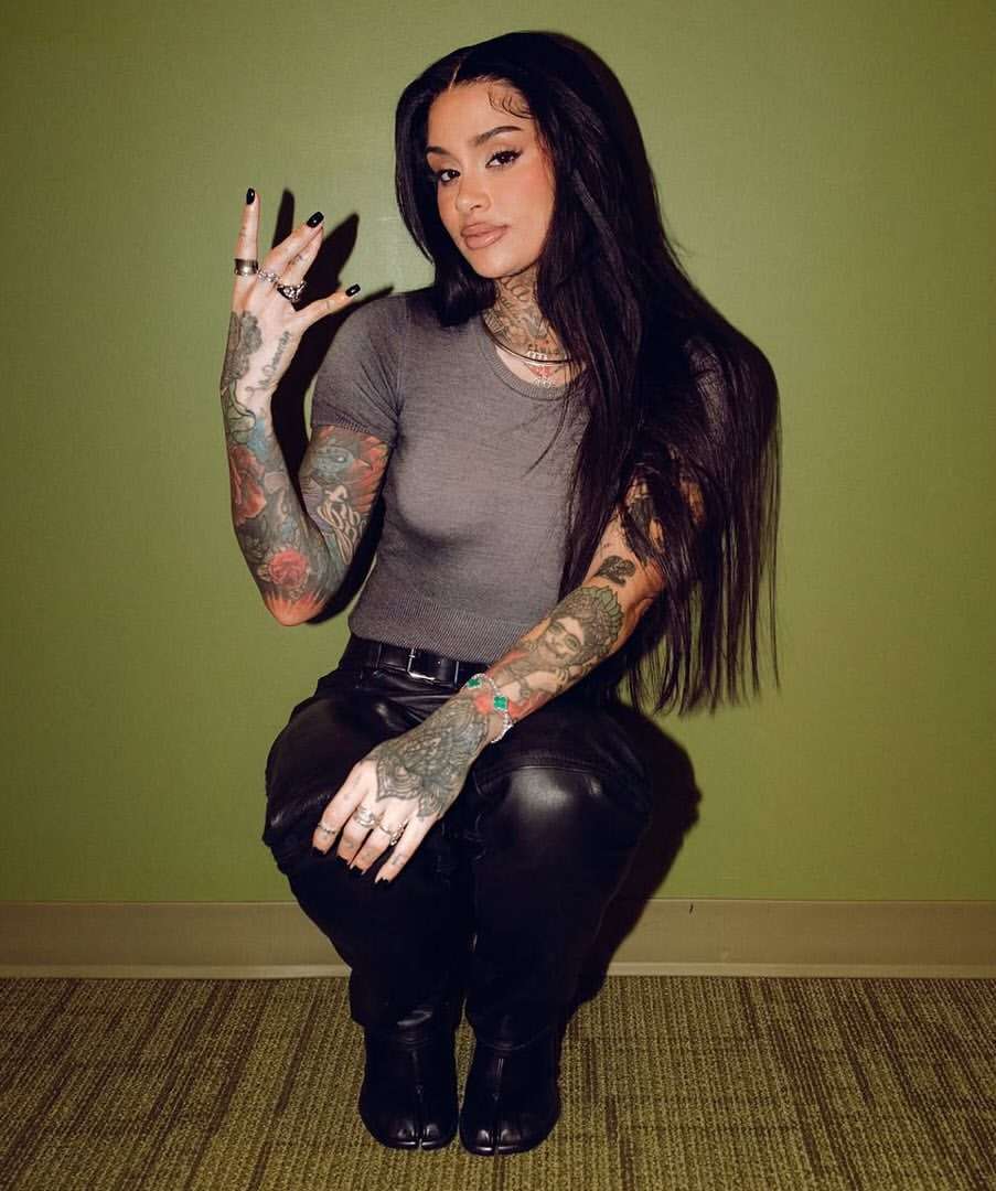Woman with long dark hair and extensive arm tattoos squatting against a green wall wearing a grey t-shirt and black pants.