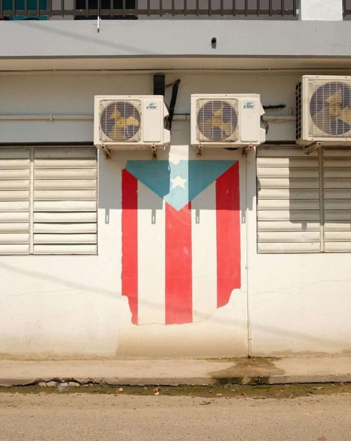 Building exterior features an air conditioner cluster above a colorful Puerto Rican flag mural, set against a neutral urban backdrop.