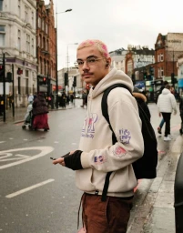 Young man with dyed pink hair and glasses standing on a wet London street wearing a hoodie and backpack.