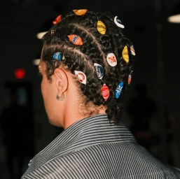Close-up portrait of a man with braided hair decorated with colorful bottle caps, wearing a striped shirt by Junior Butler