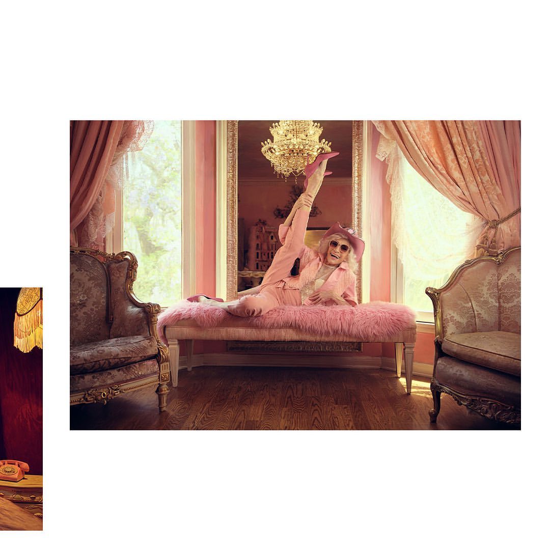 Mature woman in pink suit and hat lounges on pink chaise lounge in ornate interior with chandelier, curtains, and decorative furniture.