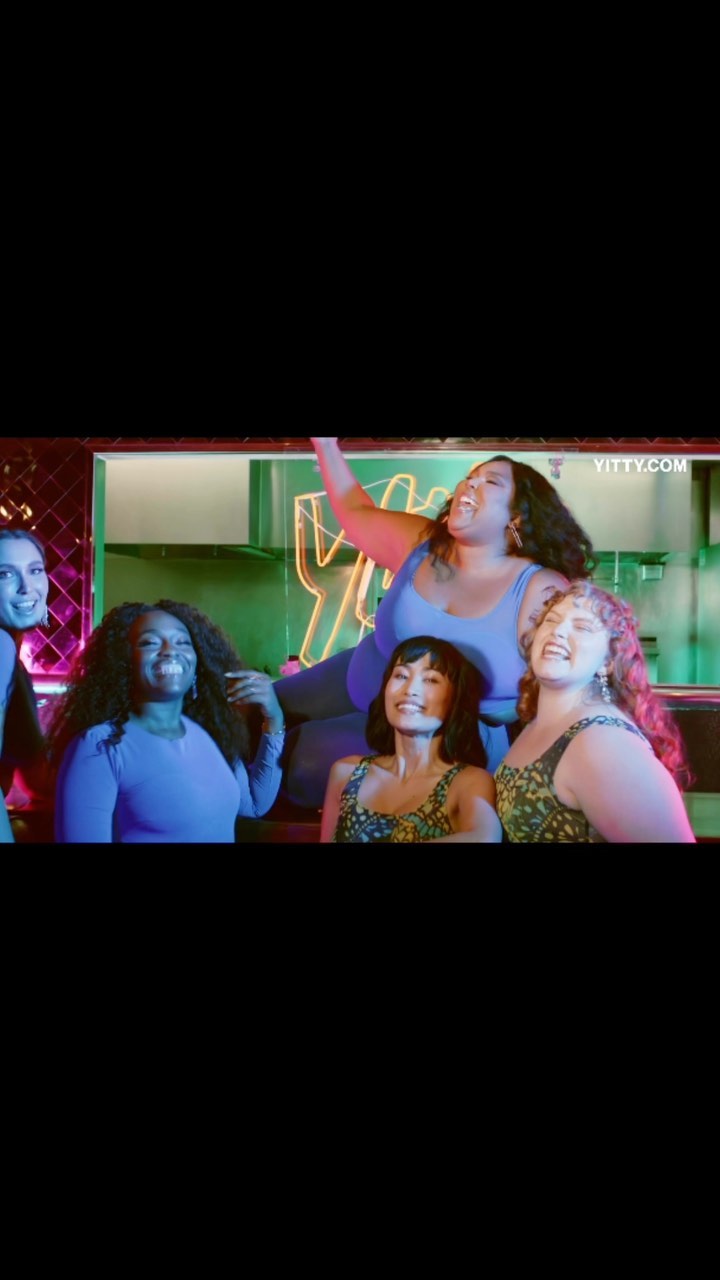 Group portrait of six women of diverse ethnicities in an indoor setting with a green neon sign in the background.