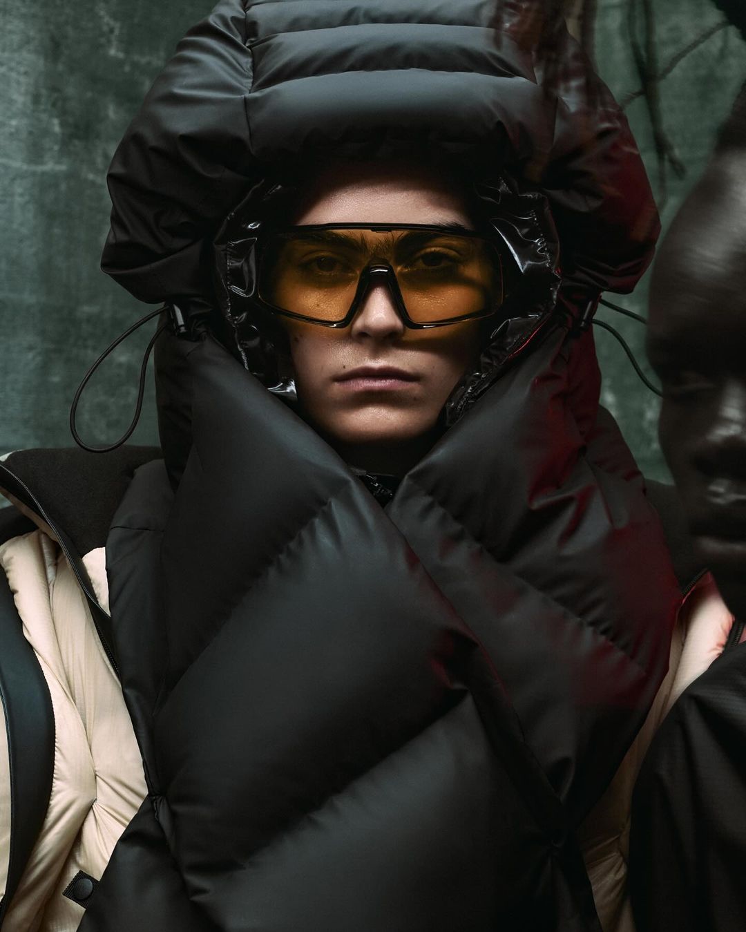 Close-up of a person with yellow tinted glasses wearing a black puffy coat and hood standing next to a dark-skinned person.