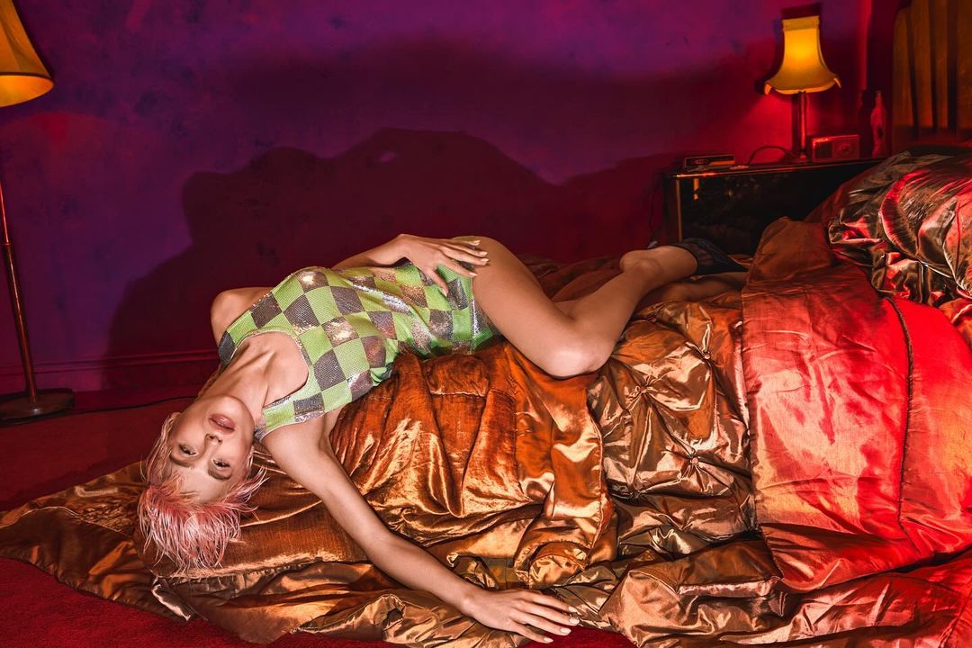 Woman with pink hair poses on a gold bed in a checkered green dress, illuminated by bedside lamp and floor lamp.