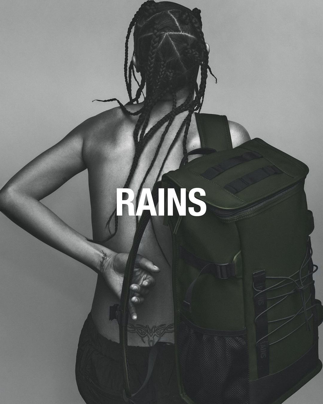 An adult female model wearing pants is seen from the back with her braids, tattoos, and a green backpack against a grey background.
