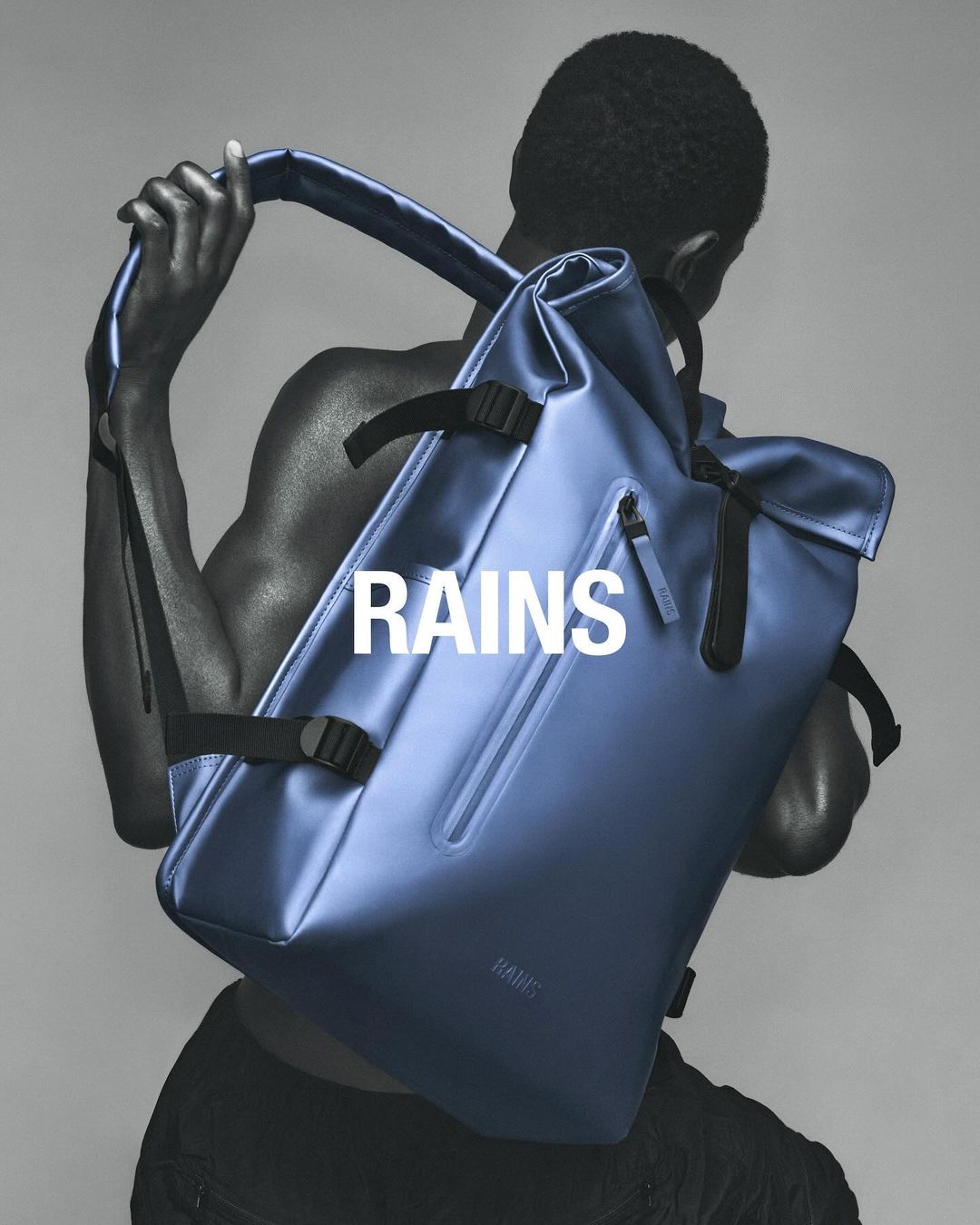 Model presents blue RAINS rolltop backpack with black straps. Dark-skinned male, shot from behind, gray studio background, simple product shot.
