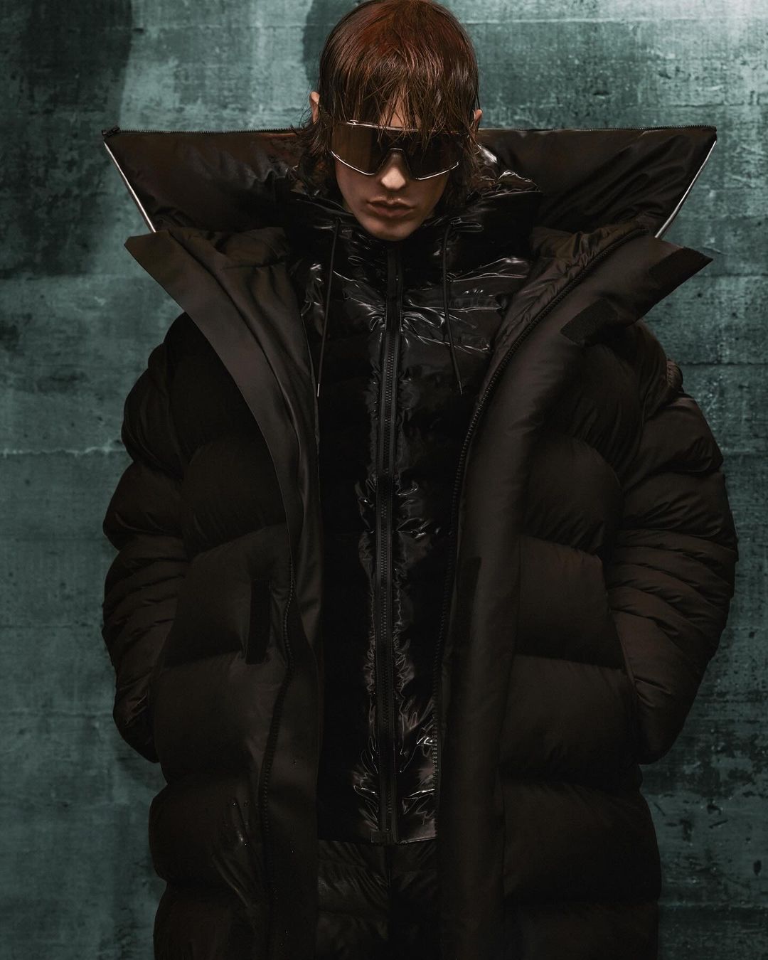 Adult model wears oversized black puffer coat and dark sunglasses in a studio shot with textured, mottled dark green backdrop.