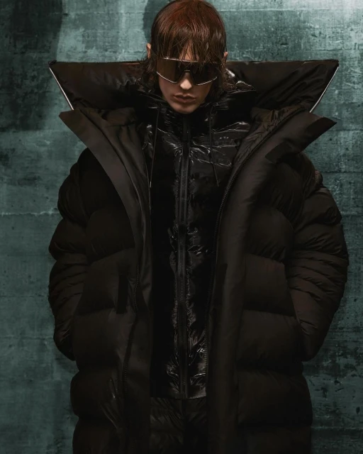 Adult model wears oversized black puffer coat and dark sunglasses in a studio shot with textured, mottled dark green backdrop.