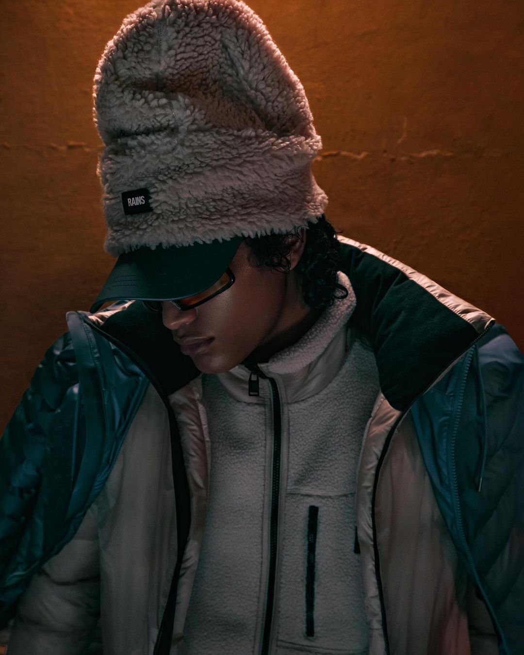 Person in beige fleece hat and jacket, with a baseball cap, posing indoors against a warm background. Fashion shot.