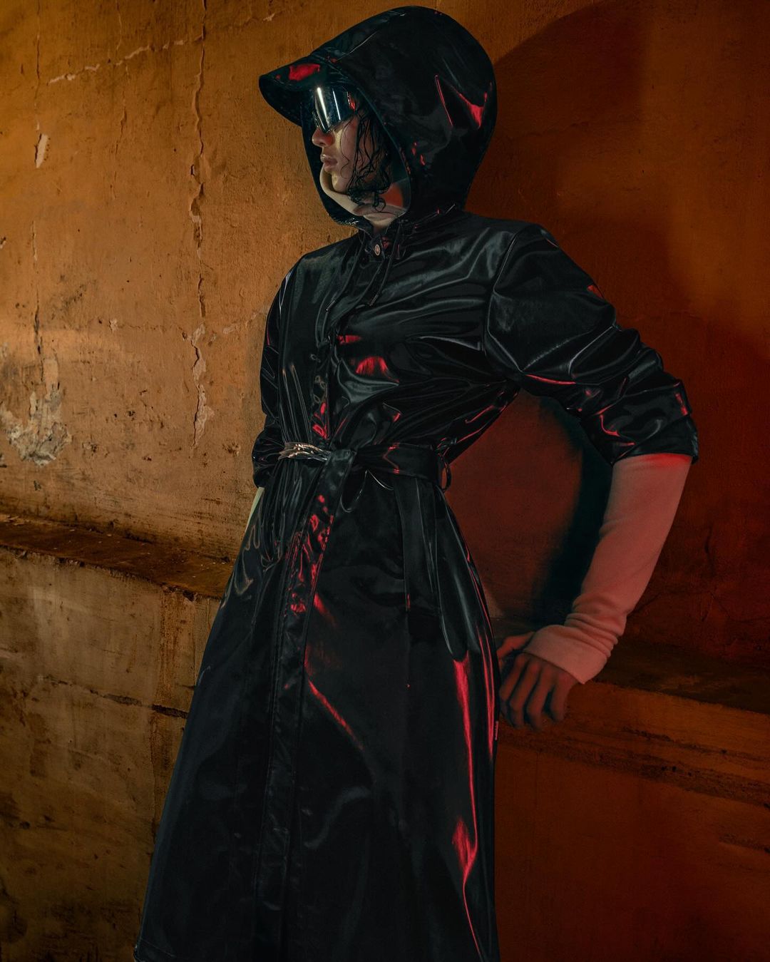 A person in a hooded black coat and futuristic eyewear stands in front of a textured orange wall with red lighting accents, dark and mysterious.