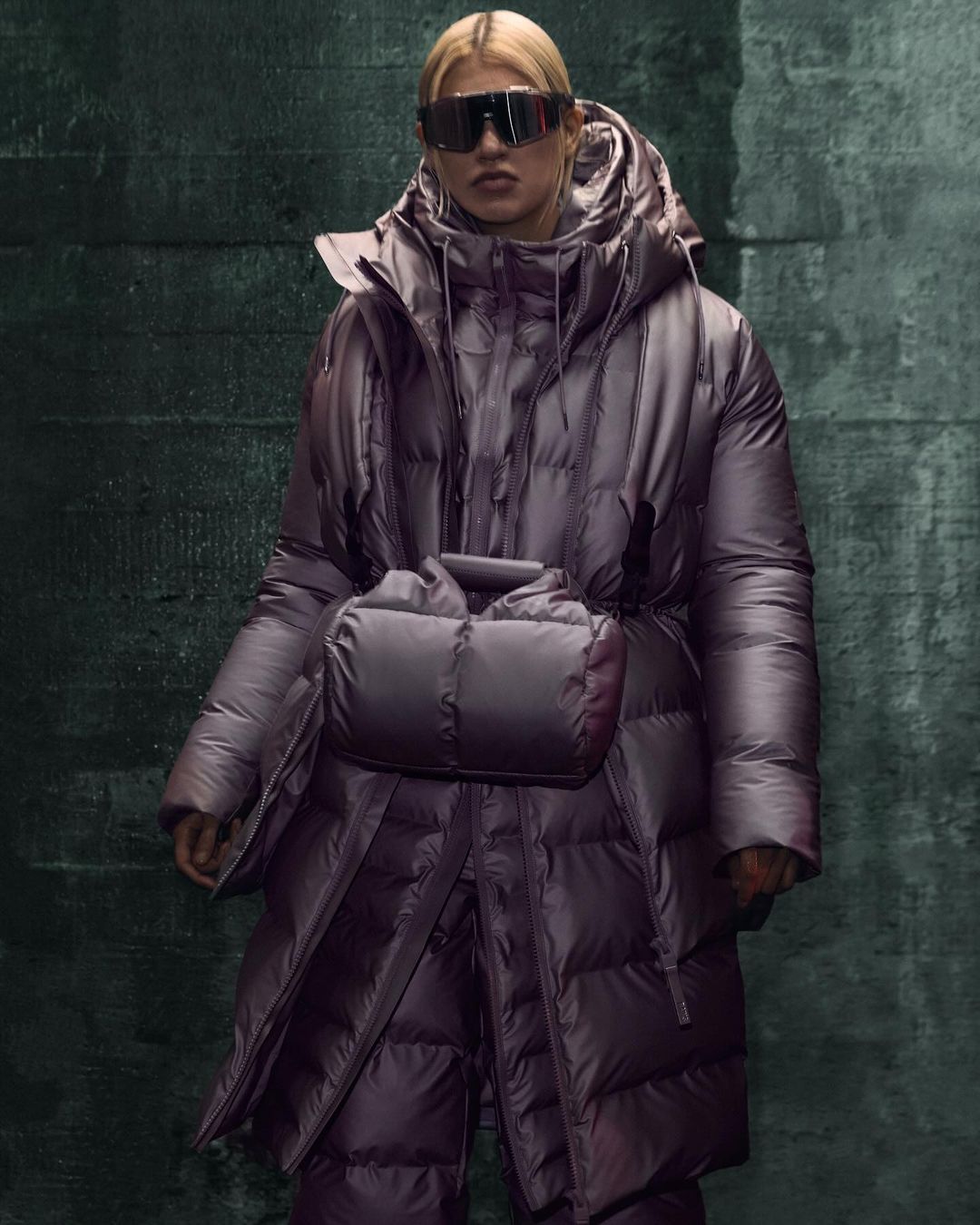 Woman in a shiny purple puffer coat and futuristic sunglasses stands against a dark grunge wall with geometric patterns in a studio shot.