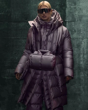 Woman in a shiny purple puffer coat and futuristic sunglasses stands against a dark grunge wall with geometric patterns in a studio shot.