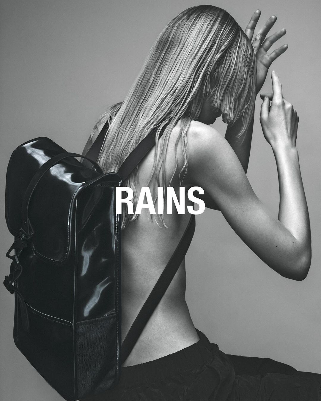 Back view of a blonde adult female model wearing a black backpack, in a studio setting with grey backdrop. Fashion photography.