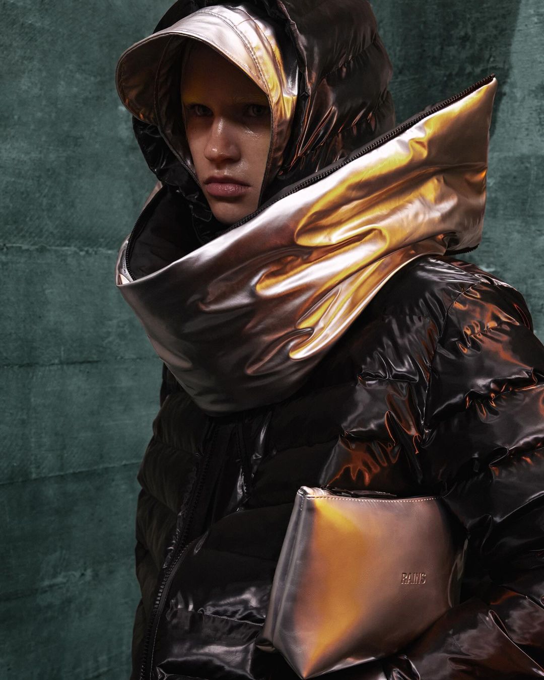 Woman in a black puffy jacket with a metallic gold hood and purse, posing in front of a dark green textured backdrop, fashion shot.