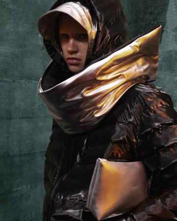 Woman in a black puffy jacket with a metallic gold hood and purse, posing in front of a dark green textured backdrop, fashion shot.