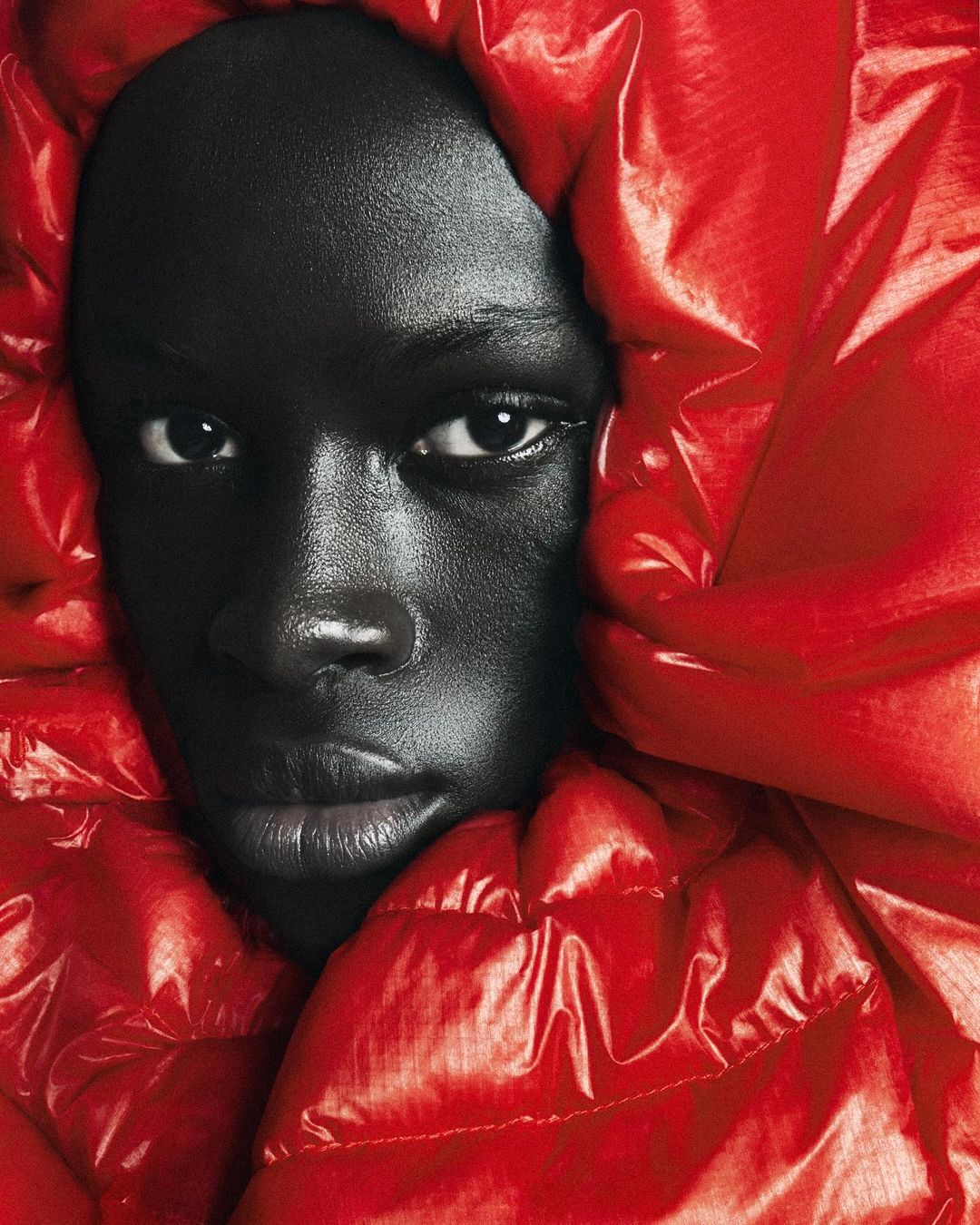 Close-up studio shot of an adult Black woman wearing a red puffy coat with a hood. Dramatic lighting highlights facial features.
