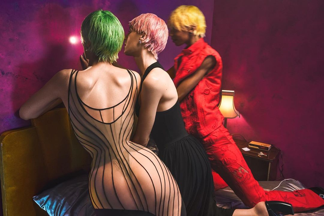 Three people with vibrant green, pink, and yellow hair posing in a bedroom setting with colorful walls and decorative lamp, shot at eye level.