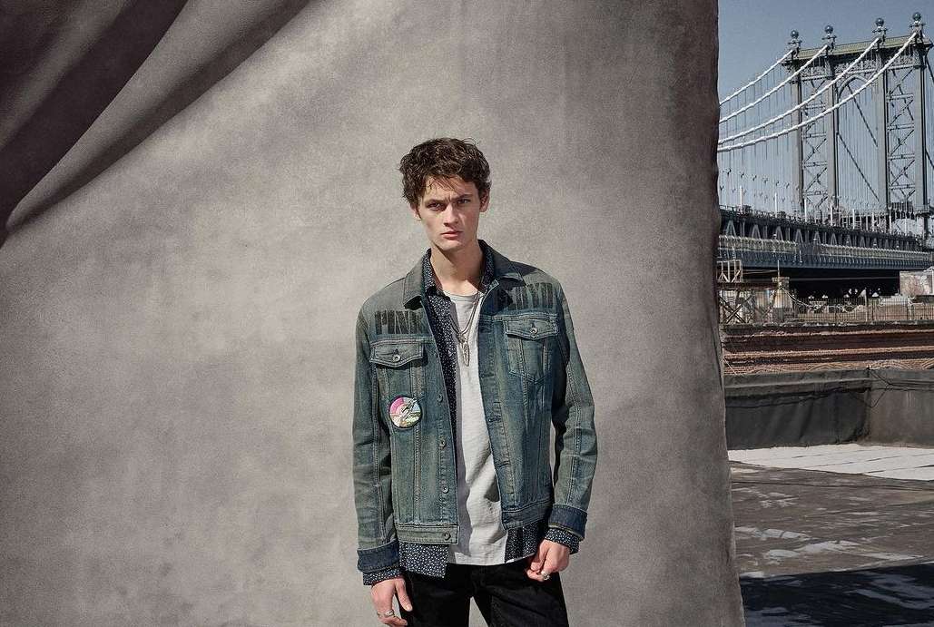 A young man in a denim jacket poses against a textured grey backdrop with a bridge visible in the background, fashion shoot.