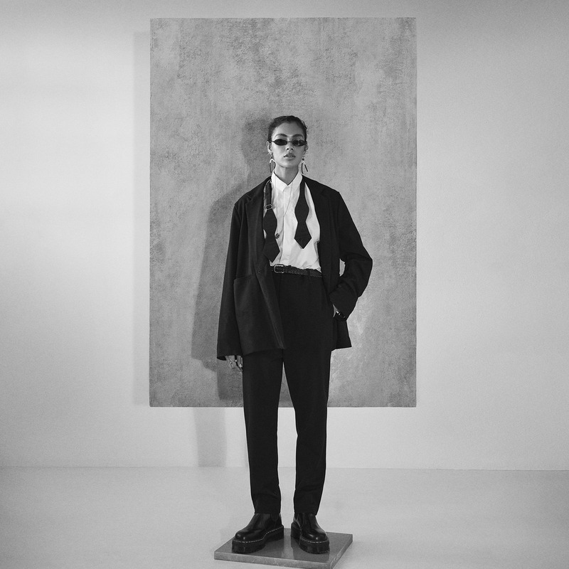 A female fashion model in a dark suit and platform shoes poses in front of a textured backdrop in a studio environment. Black and white.