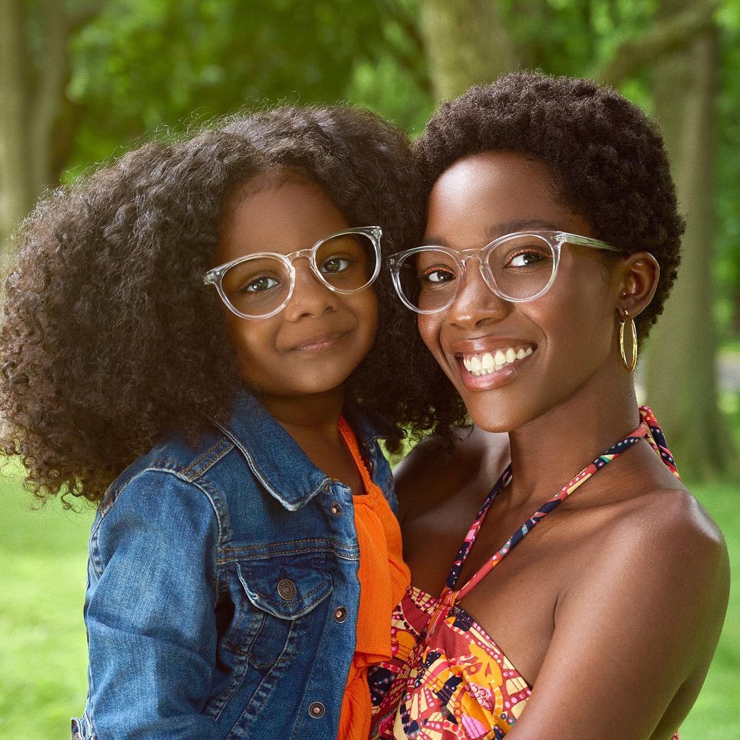 Mother and daughter wear matching clear-framed glasses and smile outdoors, with the green trees in the blurred background.