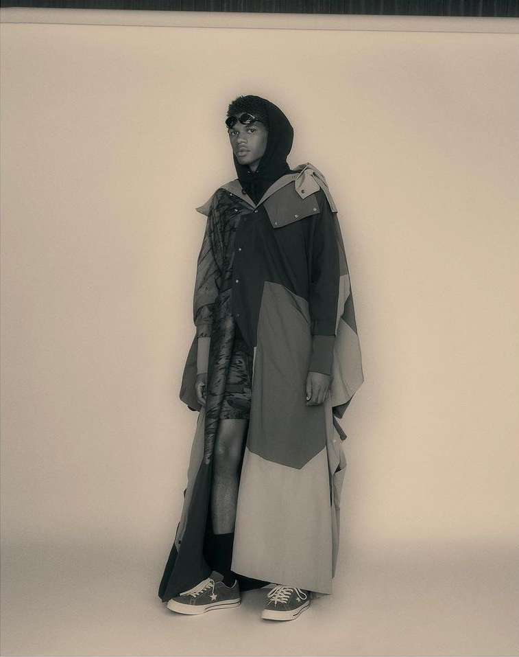 A person stands in a studio with a neutral background wearing a multi-panel overcoat, hood, sunglasses, and Converse sneakers in a fashion shot.